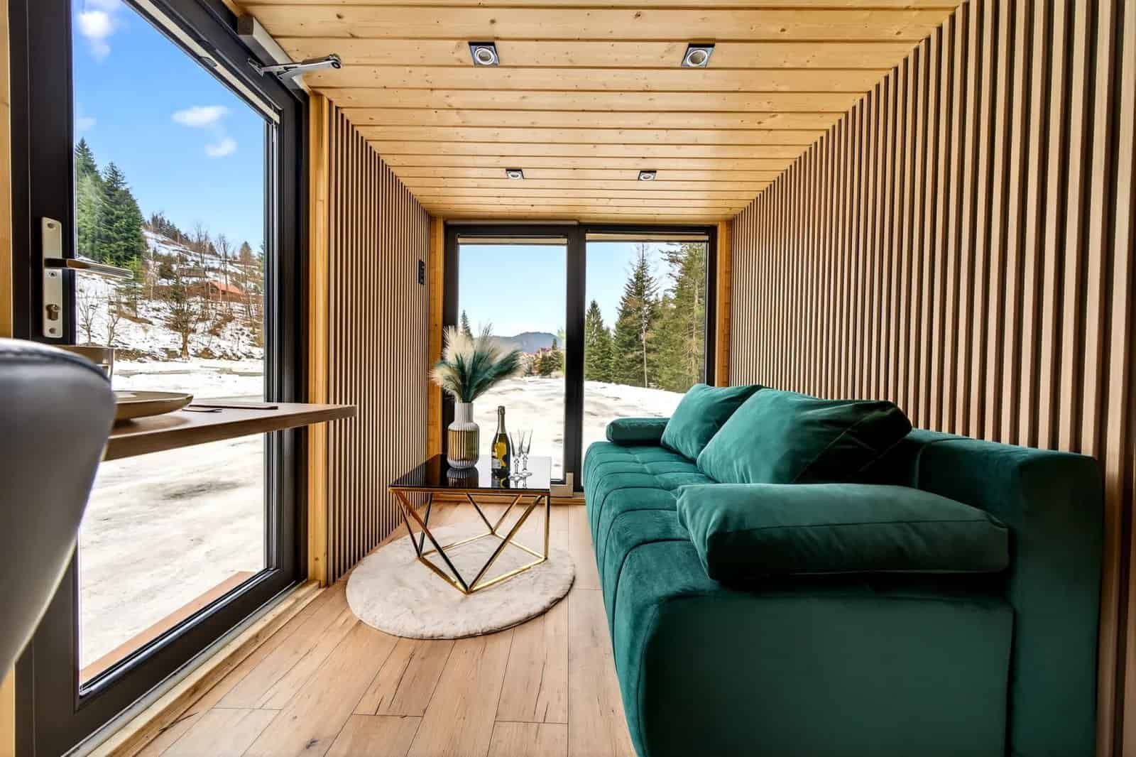 Green velvet sofa in the Tommy tiny house beside floor-to-ceiling windows framing the forest