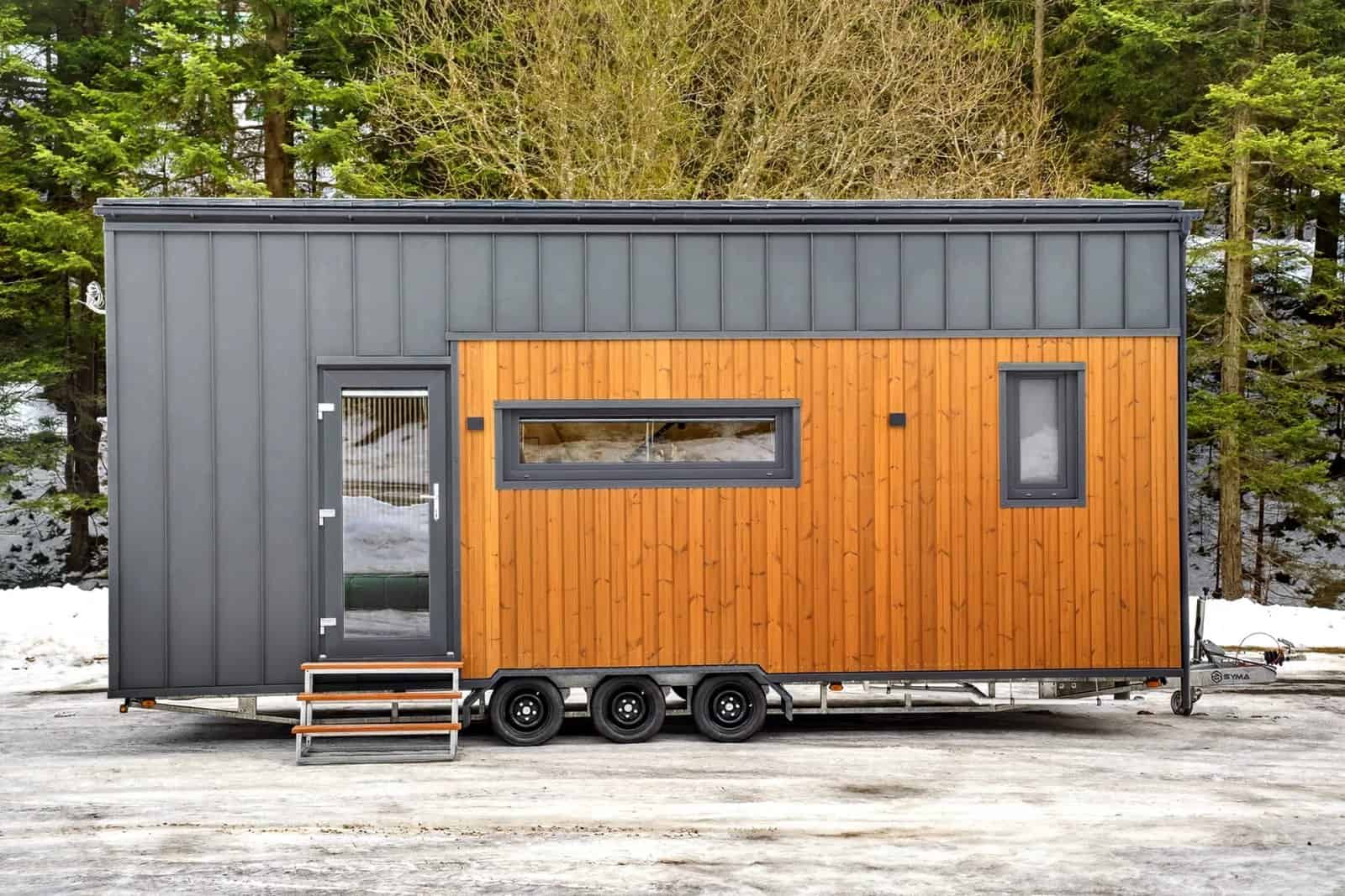 Side view of the Tommy tiny house showing the wood-clad wall, entry door, and triple-axle trailer