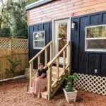 Black board-and-batten Tulsi tiny house with a cedar accent band, white-trimmed glass door, and wood steps in a fenced yard