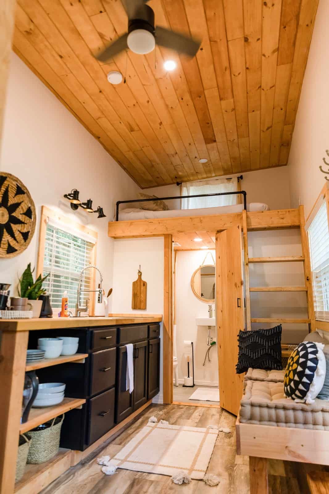 Interior of the Tulsi tiny house showing the butcher-block kitchen with black cabinets, a sleeping loft with ladder, the tiled bathroom, and a built-in bench