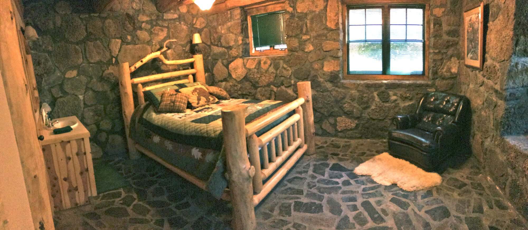 Stone-walled bedroom on the lower level with a queen log bed and leather armchair