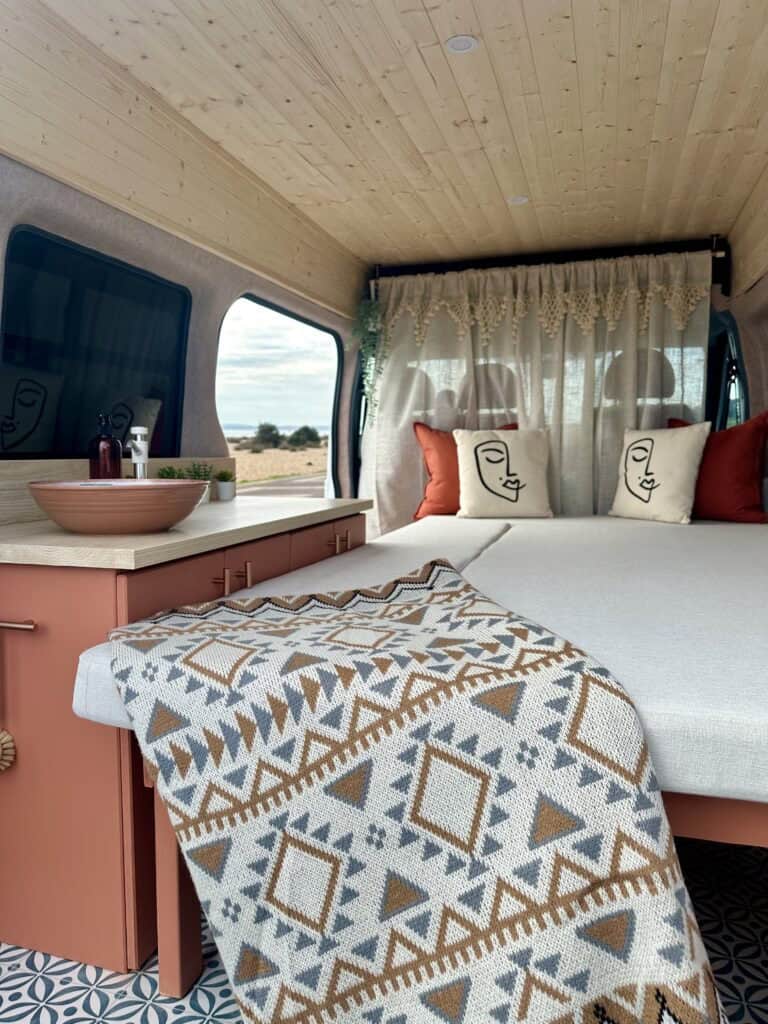 Compact van living space with a comfortable bed, decorative pillows, and a small side table. Perfect.