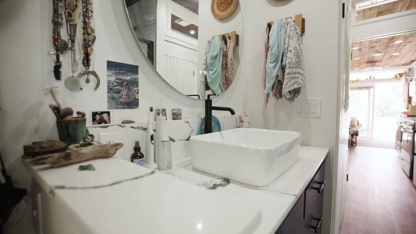 Bathroom vanity in a container home with a white vessel sink on a marble-look counter, black faucet, round backlit mirror, navy cabinets and towels on wall hooks