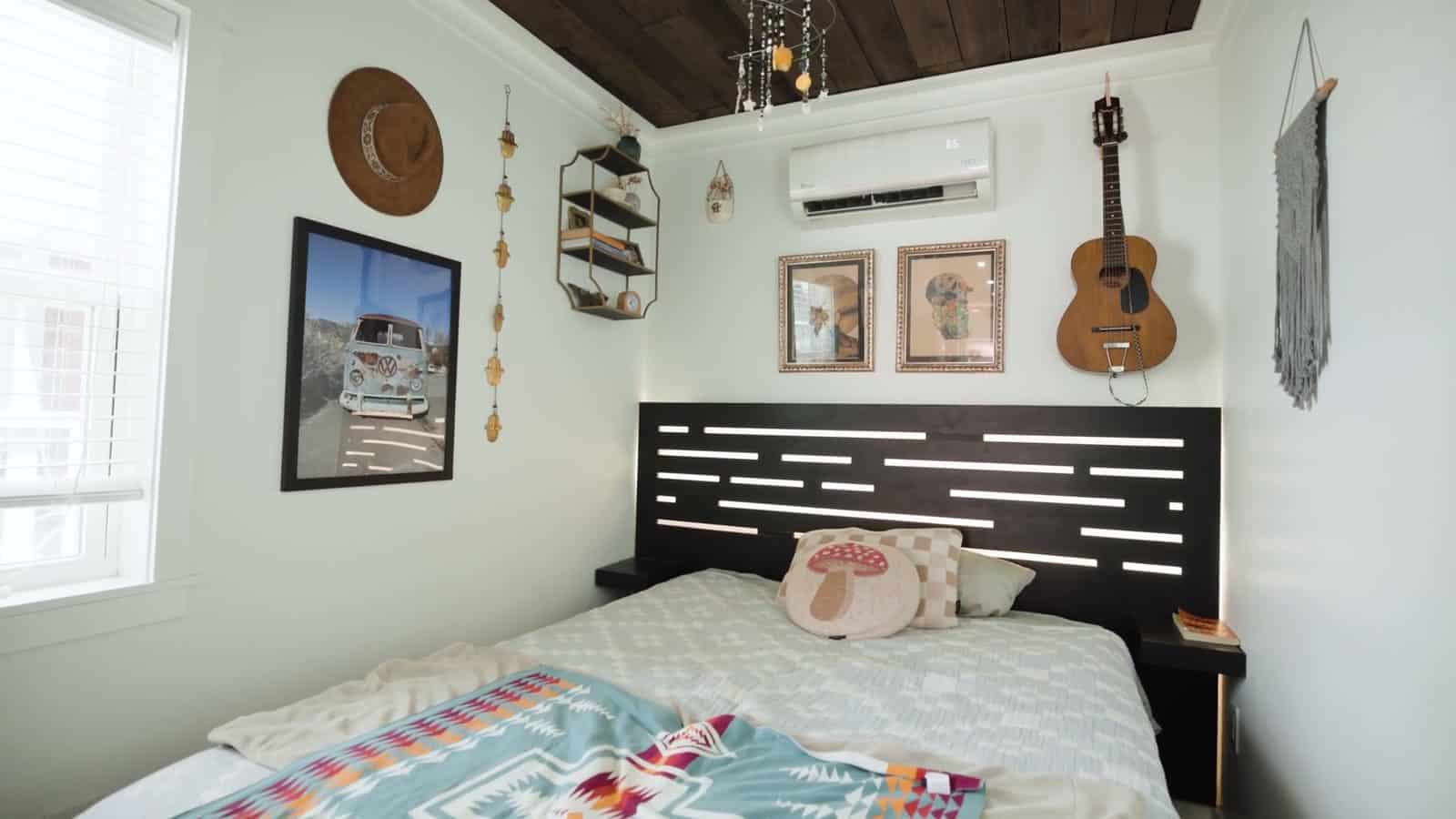 Bedroom in a container home with a queen bed under a turquoise Pendleton blanket, a black slatted headboard, mini-split, framed art, a hanging guitar and windows on both sides