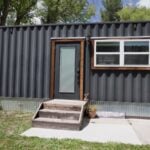 Broadside of a black 8x40 shipping container home with corrugated steel walls, a wood-trimmed entry door with wooden steps, a white-framed window, and the original container doors folded open at the end