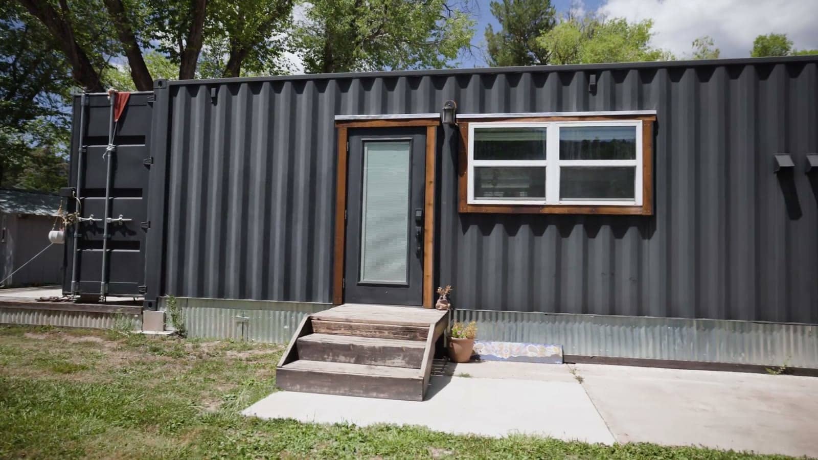 Broadside of a black 8x40 shipping container home with corrugated steel walls, a wood-trimmed entry door with wooden steps, a white-framed window, and the original container doors folded open at the end