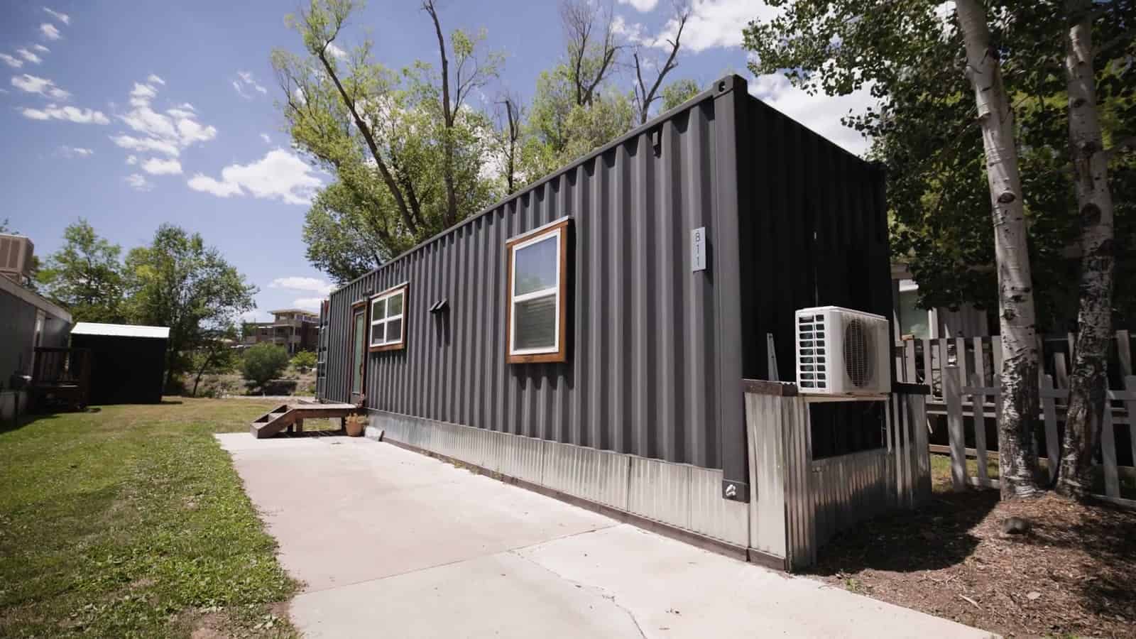 Long side of a black 8x40 shipping container home with a wood-trimmed window, a mini-split condenser and a skirted base beside a concrete driveway
