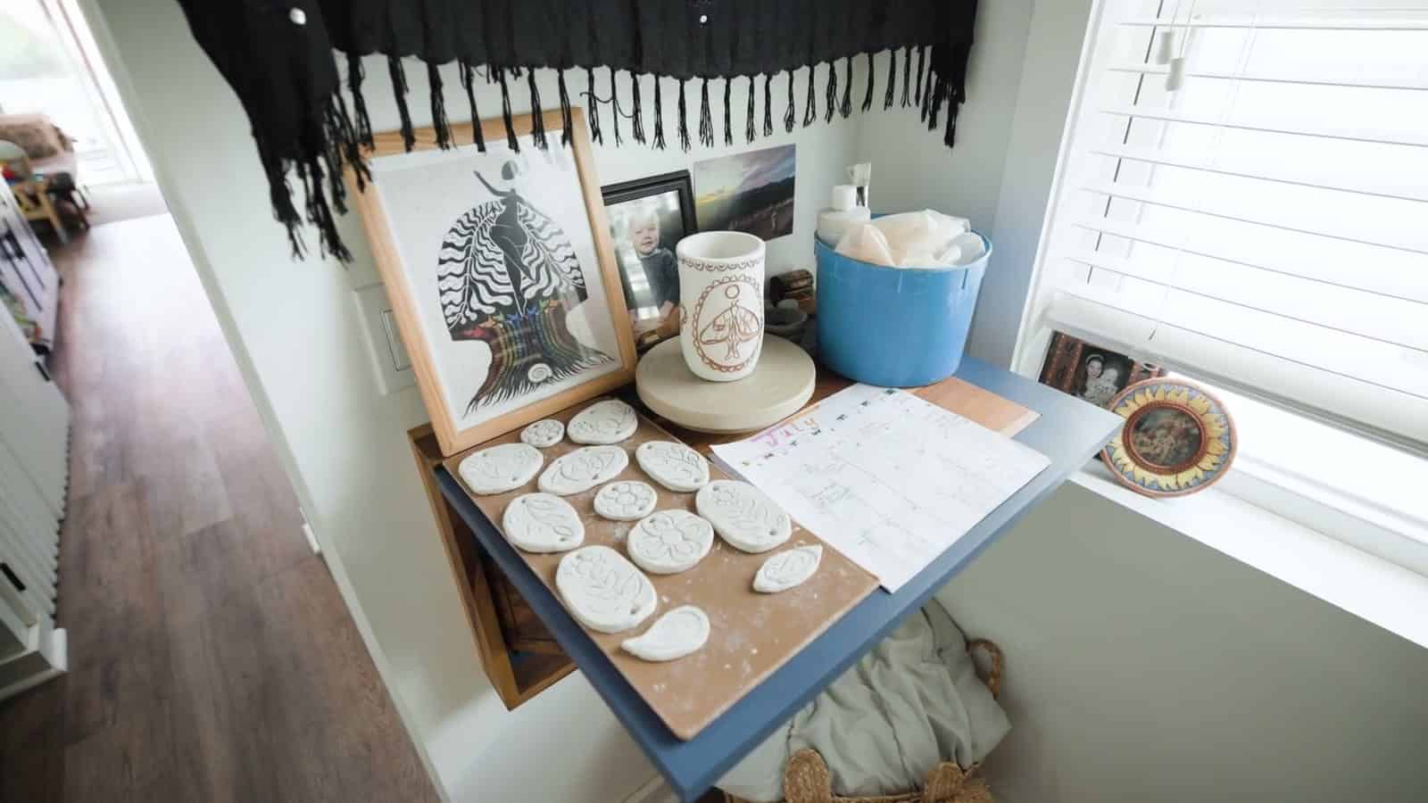 Wall-mounted fold-down desk in a container home bedroom holding pottery pieces, a framed print and supplies under a window