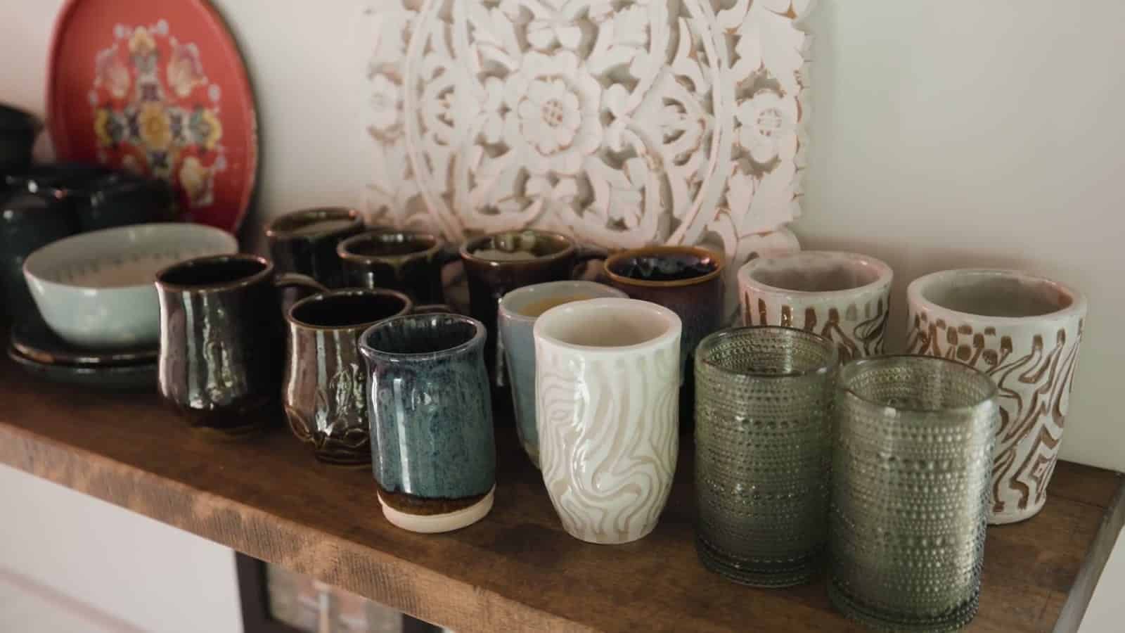 Handmade ceramic mugs and cups in blue, white and black glazes lined up on a floating wood shelf in front of a carved white panel