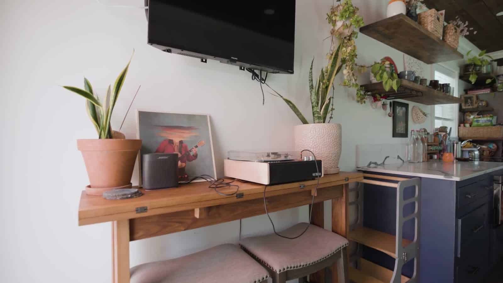 Living area of a container home with a wall-mounted TV, a narrow wood console holding a record player and plants, pink upholstered bar stools, and the navy kitchen counter beyond
