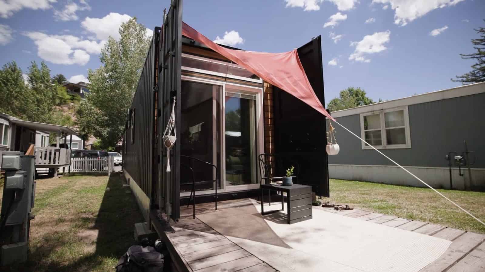 Patio end of a black 8x40 shipping container home with sliding glass doors, the original cargo doors swung open, and a coral shade sail stretched over a concrete patio