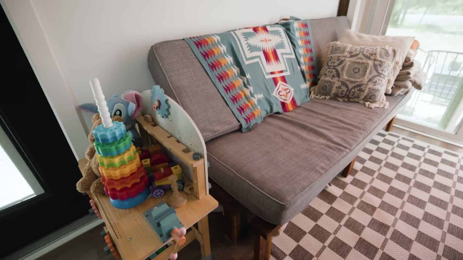 Gray pullout couch with a turquoise Pendleton-style blanket and pillows beside a window, with a stacking toy on a small shelf in front