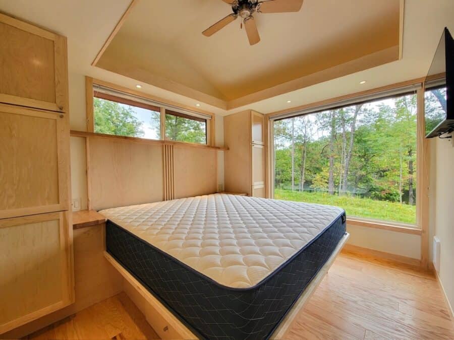 Cozy tiny house bedroom with large windows overlooking a lake and forest.