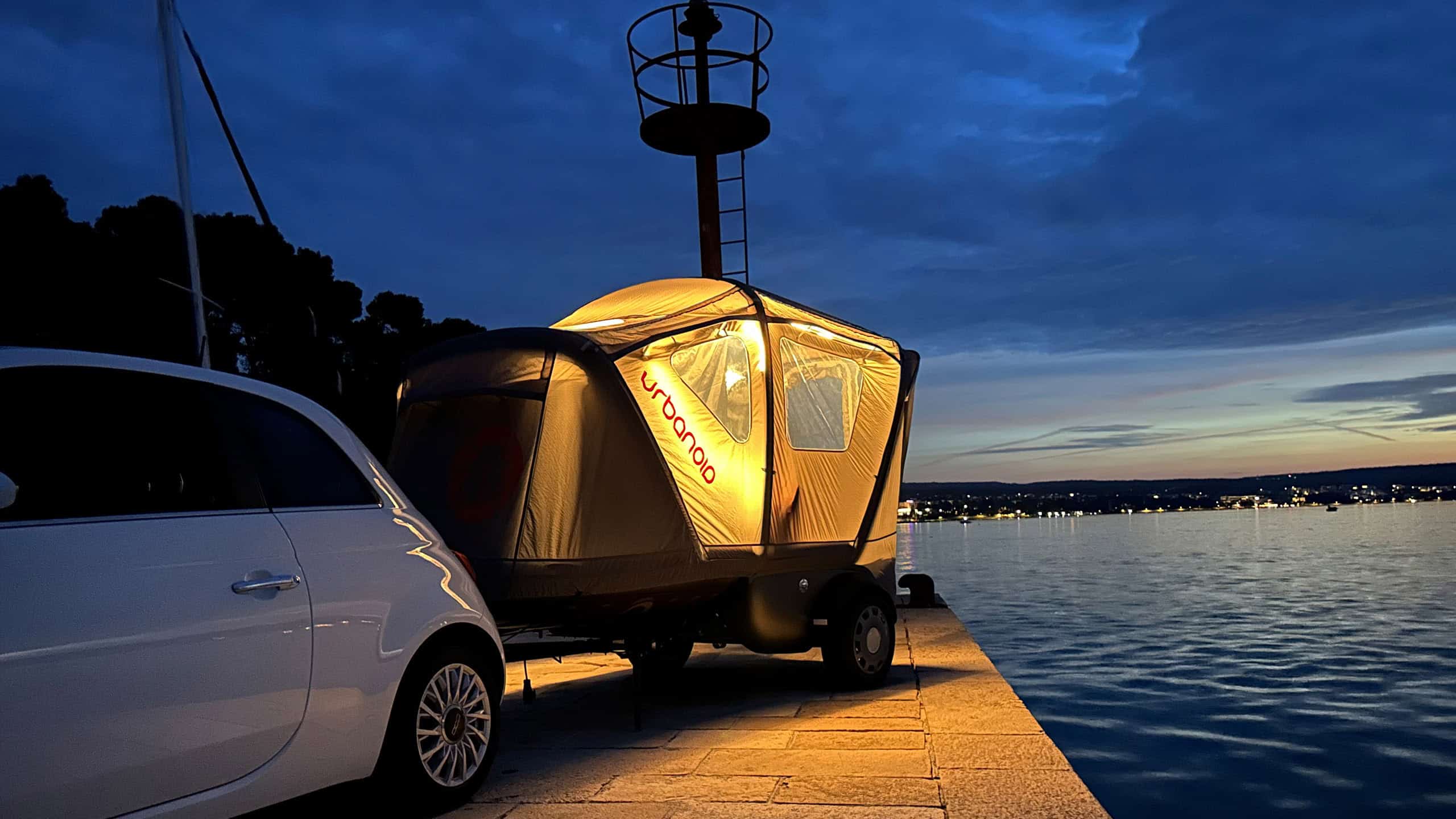 Urbanoid Booba tent glowing from interior lighting at dusk on a waterfront promenade