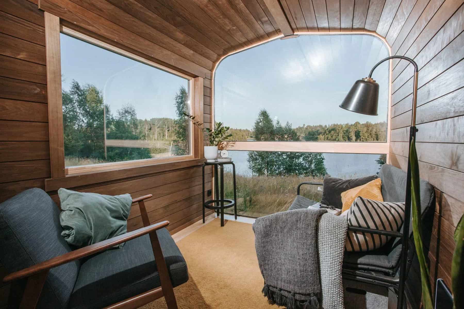 The Nature Pod set up as a lounge with a gray sofa, armchair, and floor lamp facing the panorama window and a lake