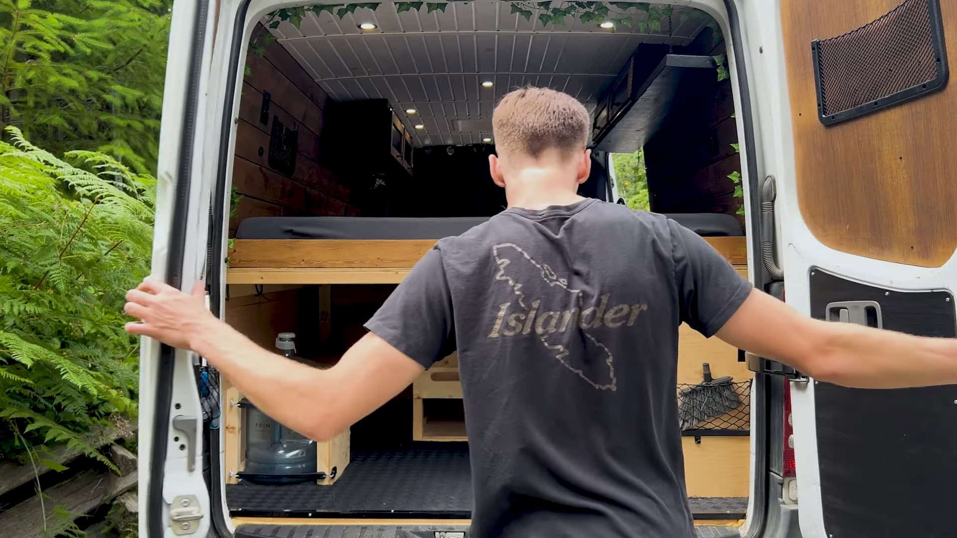 Rear doors open to a forest campsite, revealing the finished bed and wood-lined interior