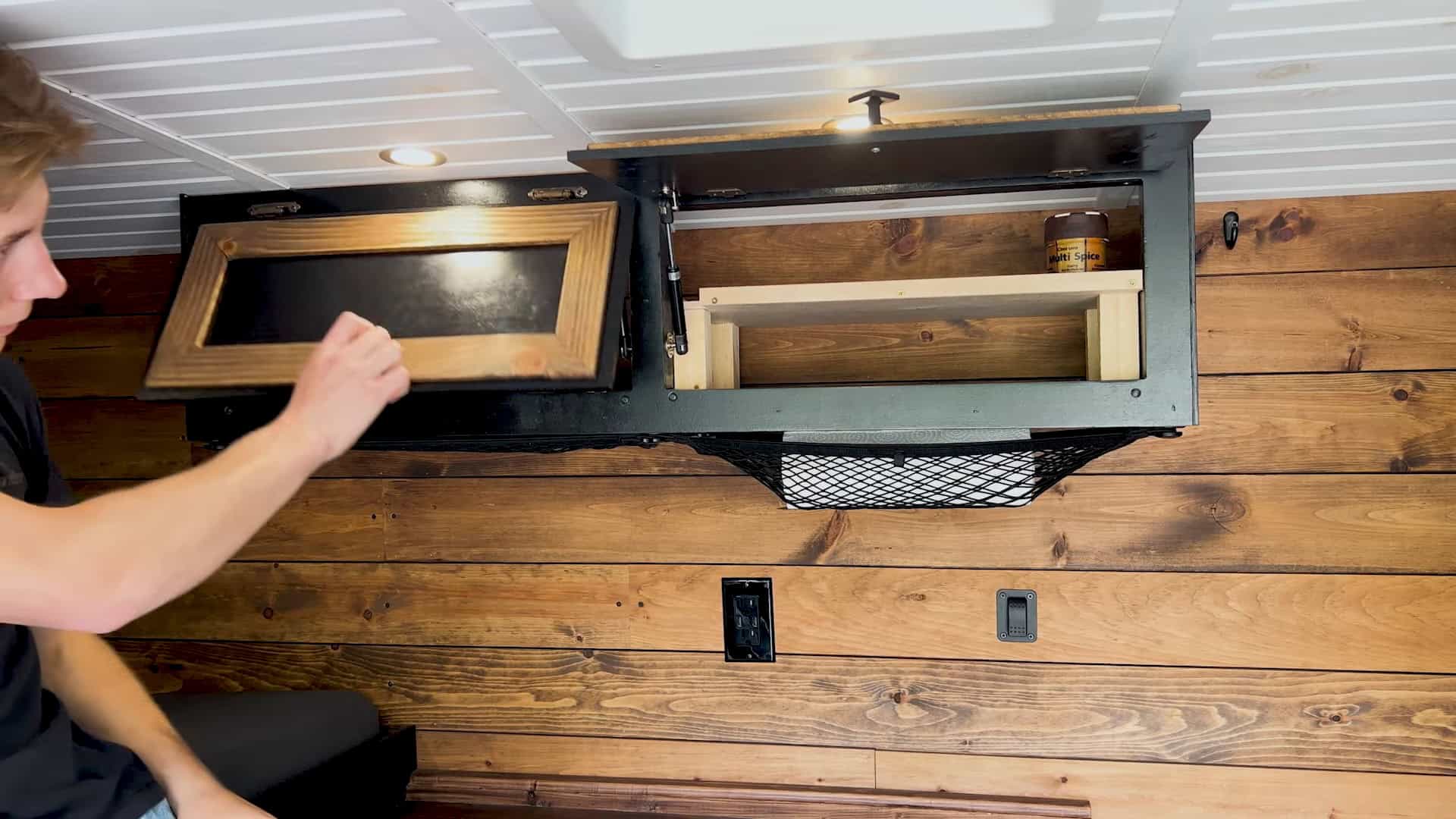 Overhead cabinet with a gas-strut door open, showing spice storage and a cargo net
