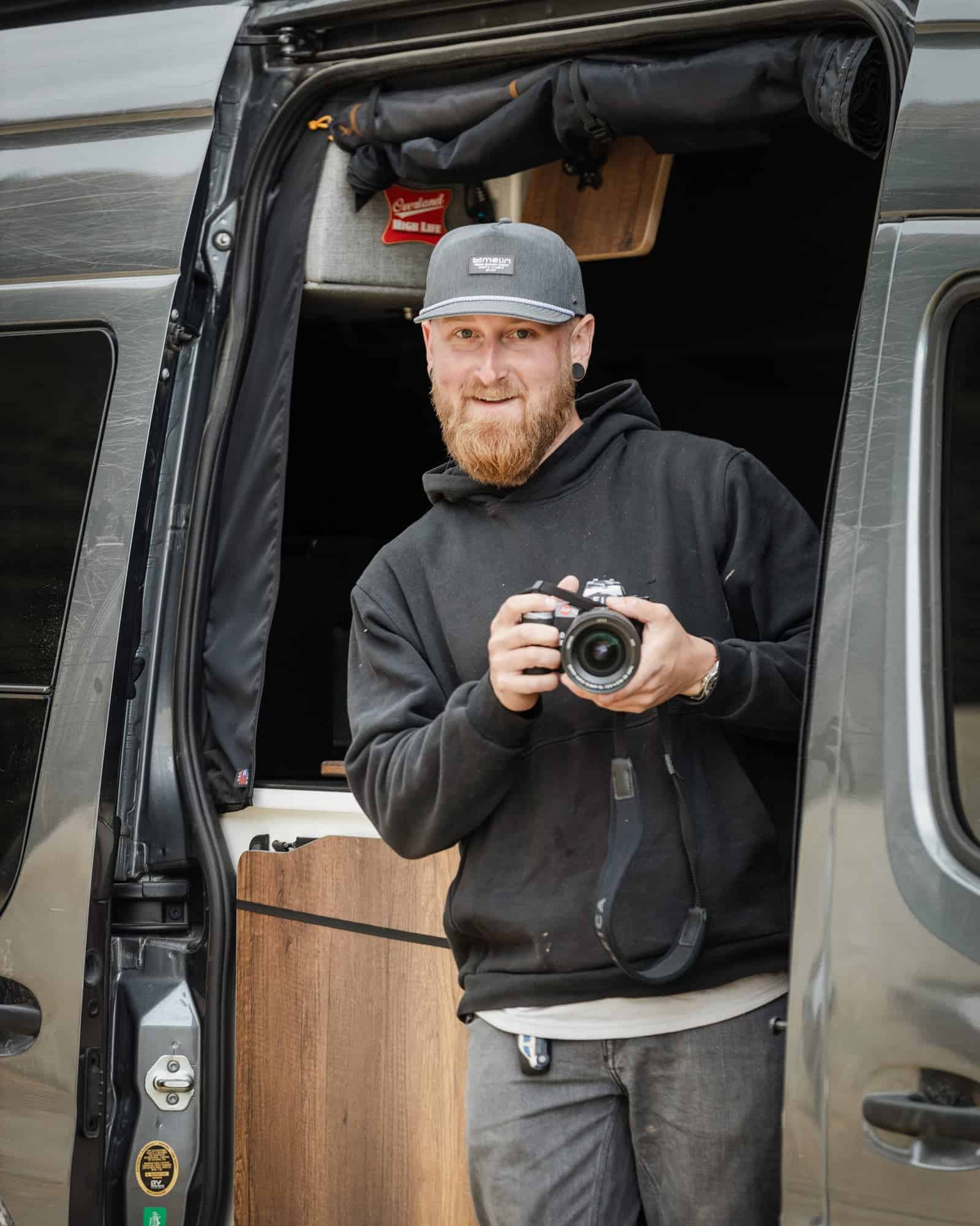 Van owner standing in the open side door of a grey adventure van holding a camera