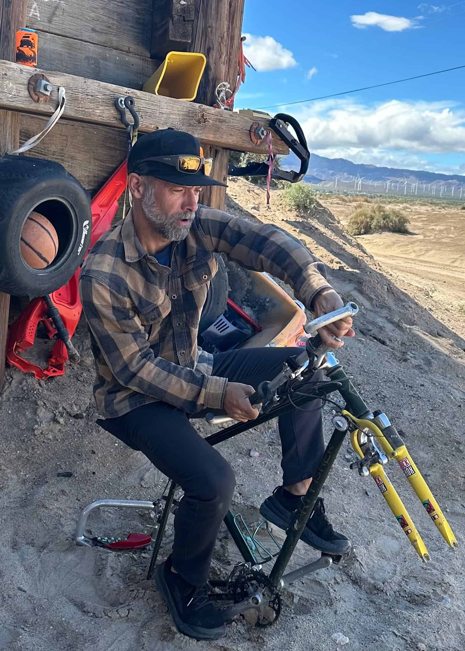 Event guide working on a mountain bike frame outdoors in the desert with wind turbines in the distance