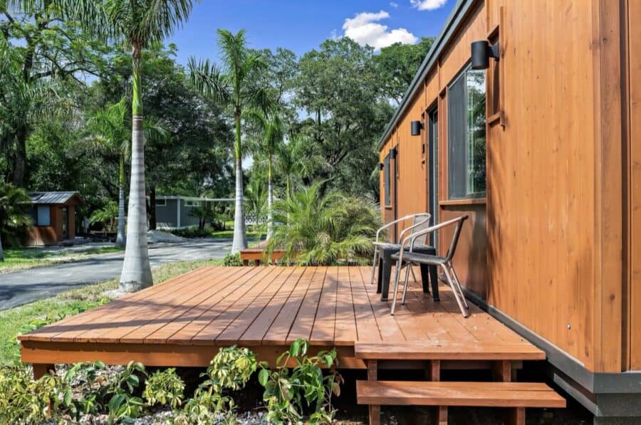 Small wooden deck attached to a tiny house with tropical plants and palm trees in the background.