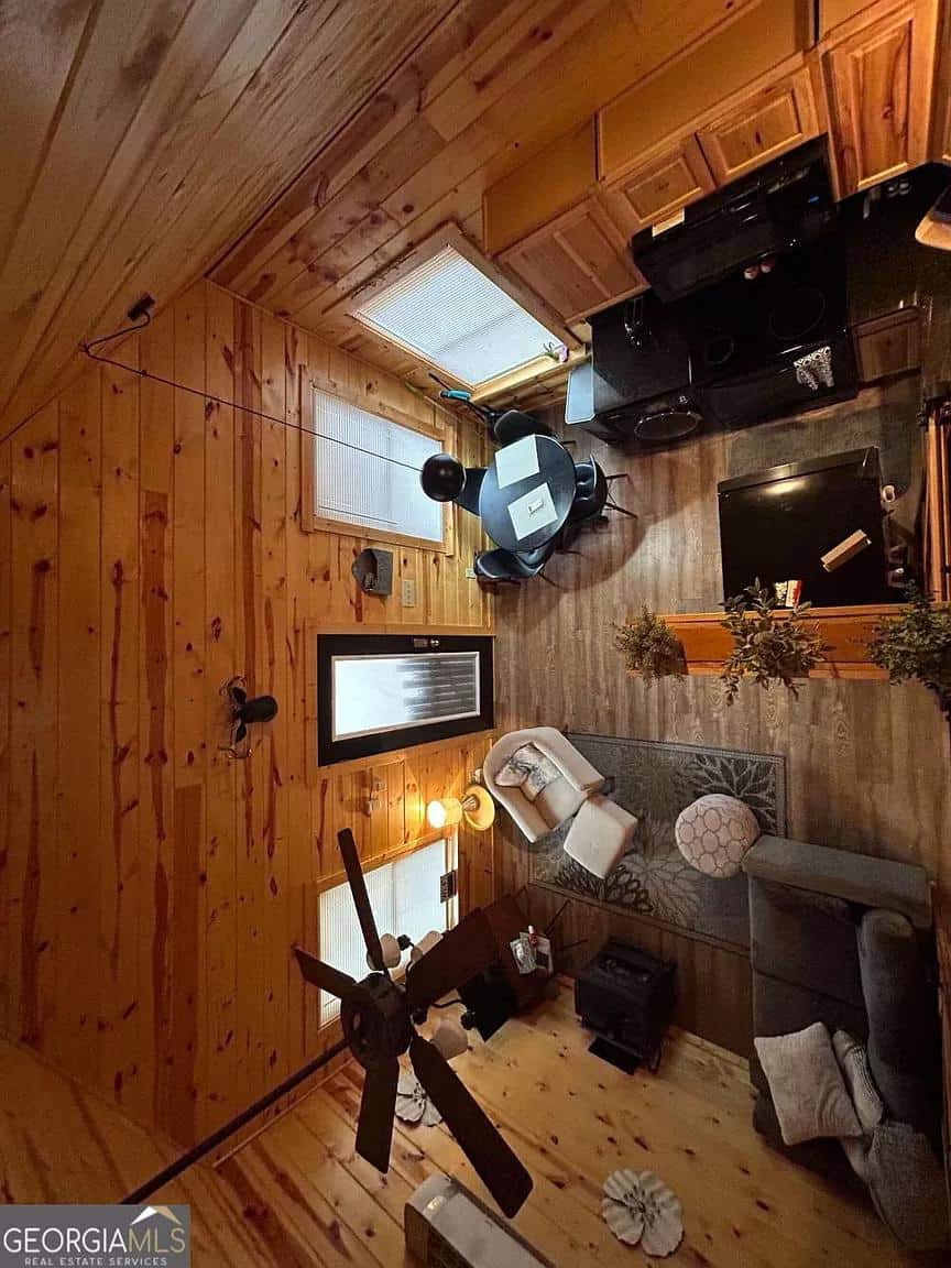 Small cabin interior with wood walls, cozy furniture, and a ceiling fan in a forest setting.