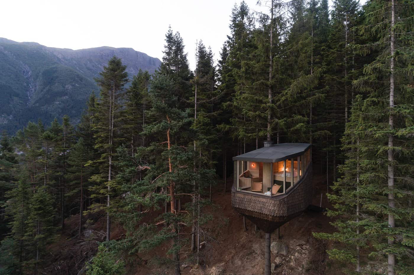 Woodnest Treehouse cabin glowing at dusk among pine trees in the Norwegian forest near Odda with mountains behind