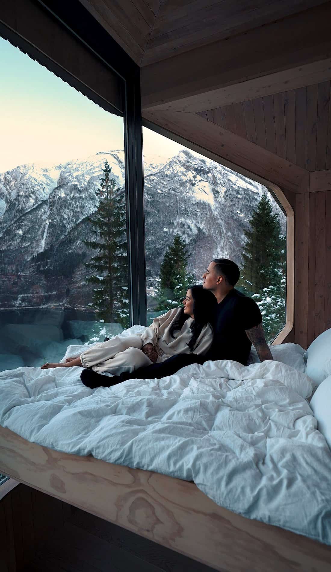 Couple on the bed inside Woodnest Treehouse looking out at snowy mountains and fjord through glass wall