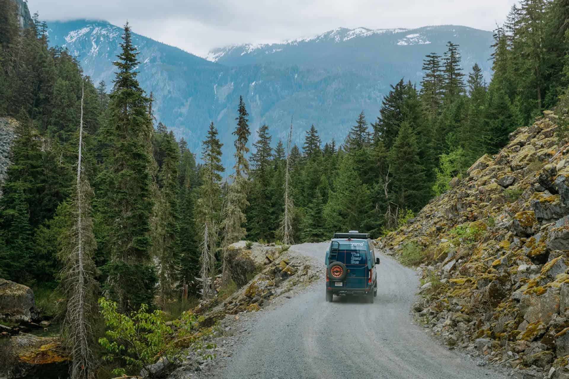 GB01 Gear Bus driving a gravel mountain road through evergreen forest with snowy peaks ahead
