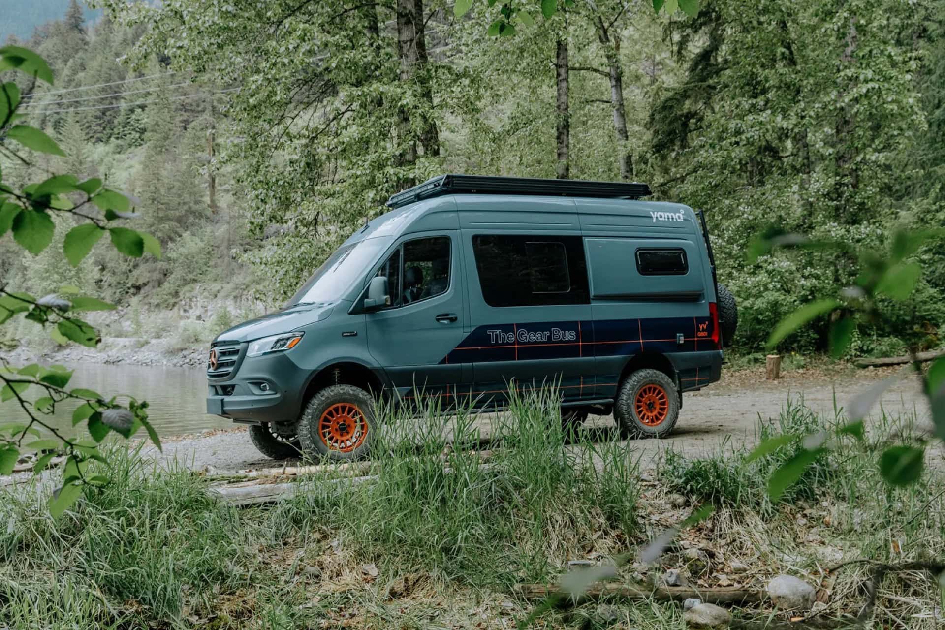 Slate-green Yama Vans Gear Bus GB01 Sprinter with orange wheels and a pop-top, parked by a river