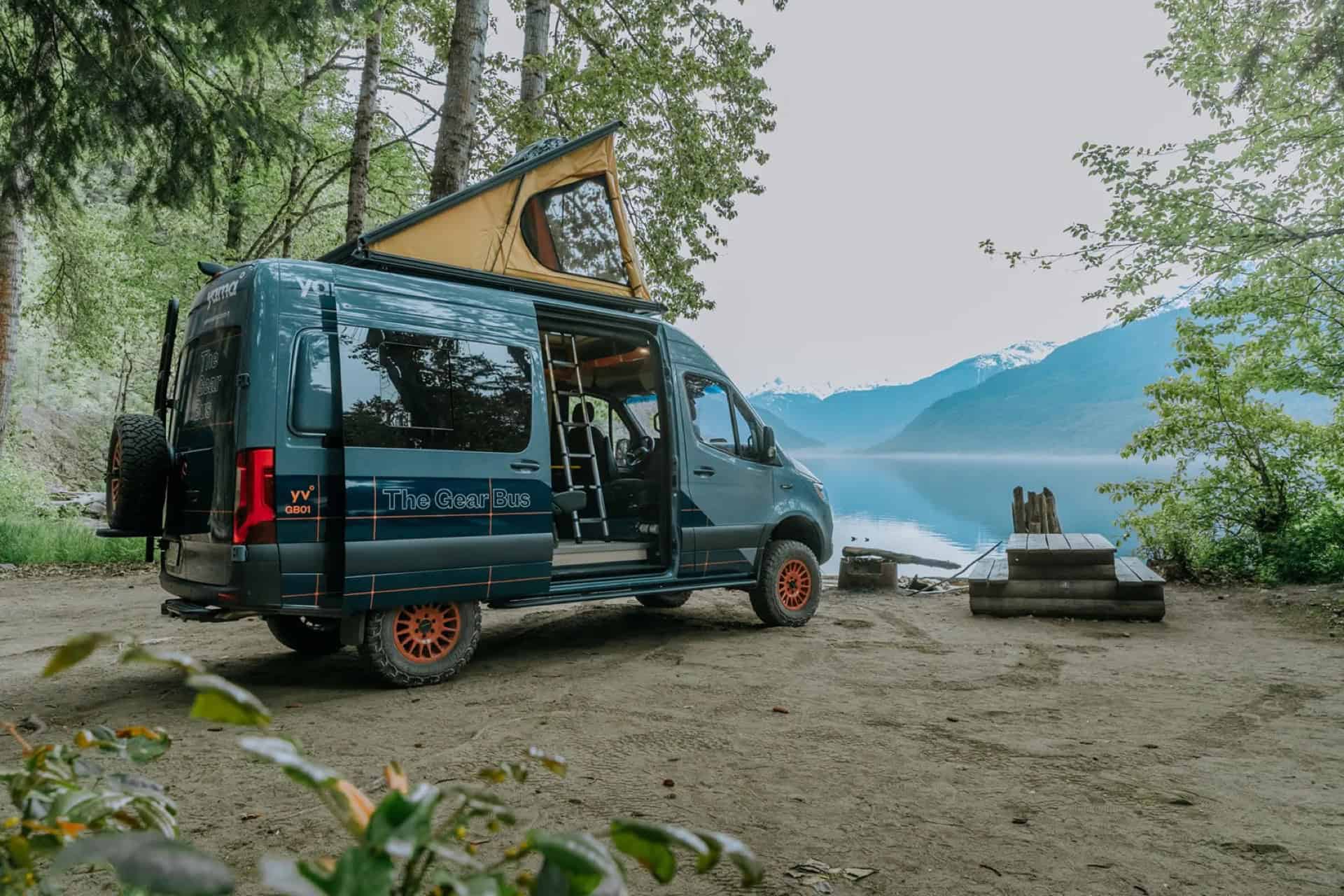 GB01 Gear Bus parked at a lakeside campsite with its pop-top raised and mountains behind