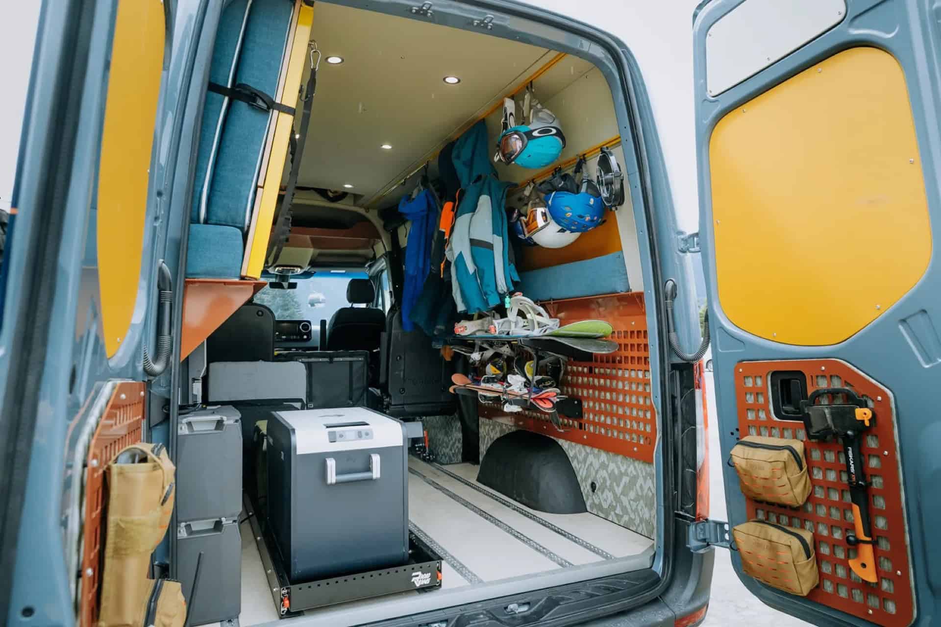 GB01 rear doors open in winter showing skis, a snowboard, jackets, helmets, and a slide-out fridge