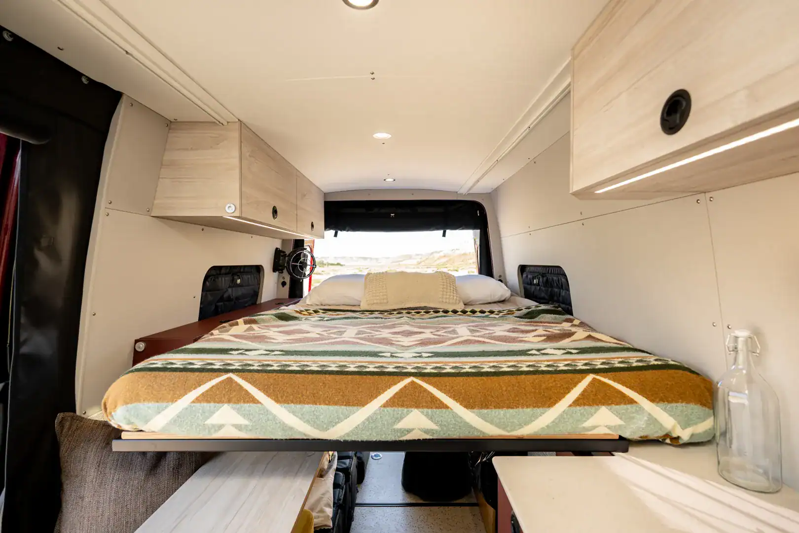 The lengthwise fixed bed in the Yama Vans Brio with a patterned blanket, maple cabinets, and a large rear window