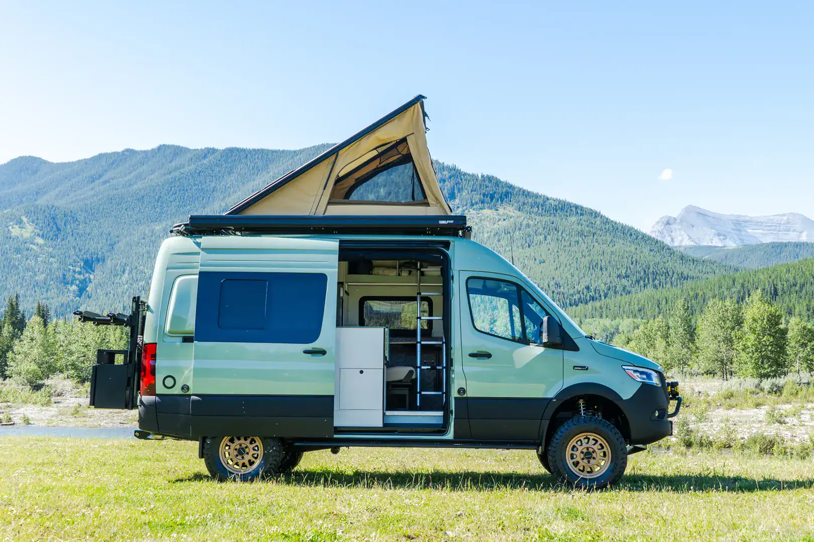 The Yama Vans Fitz, a sage-green Mercedes Sprinter 144 camper van with its pop-top raised and sliding door open in a mountain meadow