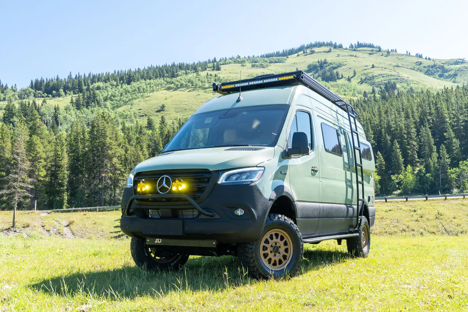 The front of the sage-green Yama Vans Fitz Sprinter 144 with a roof light bar and off-road tires in a meadow