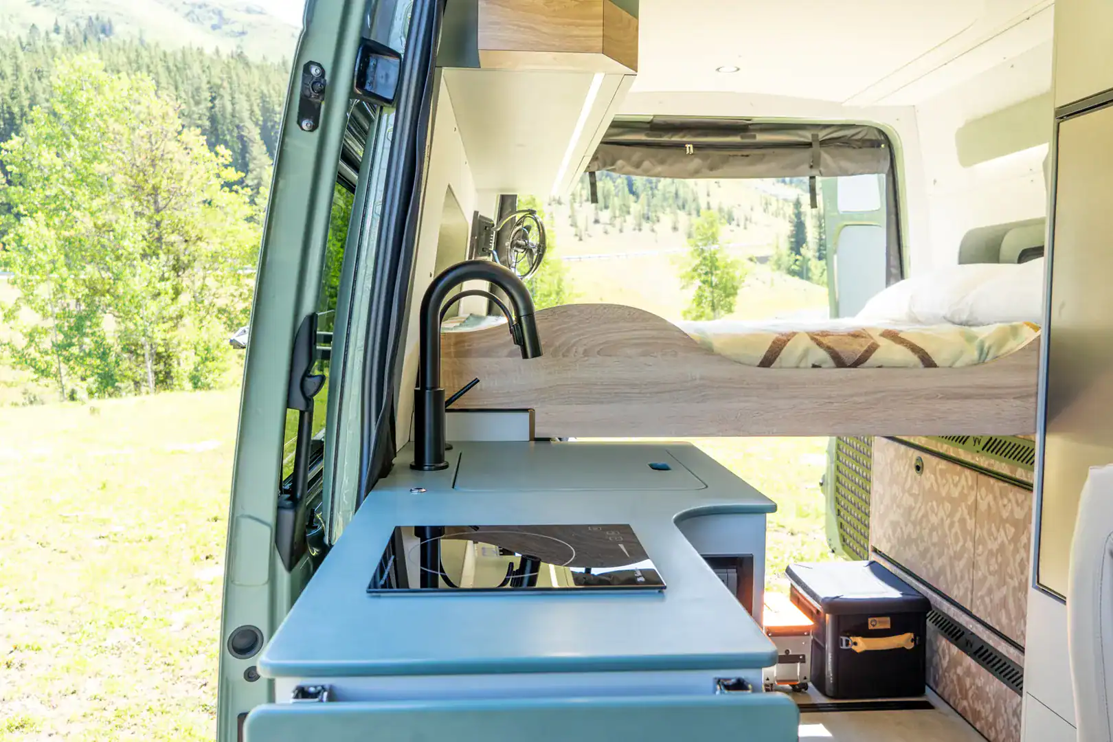 The Yama Vans Fitz galley with an induction cooktop, black faucet, and pale blue-green counter, with the bed behind
