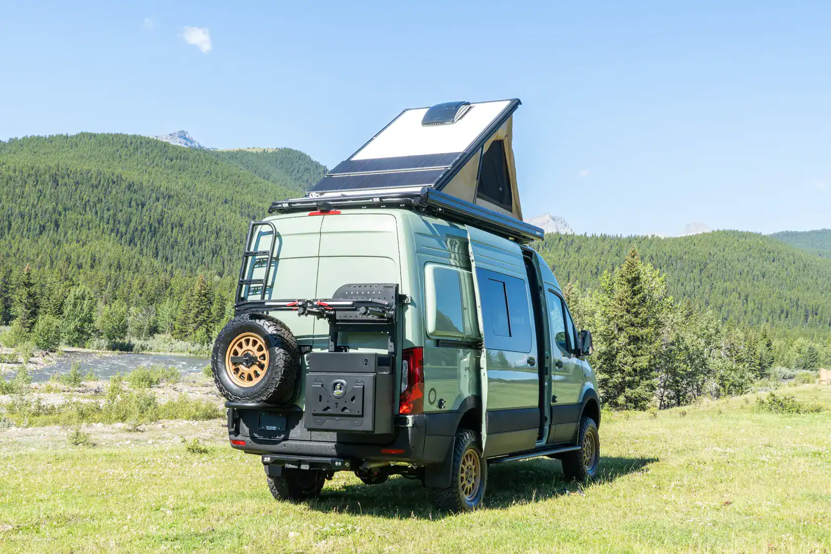 The rear of the Yama Vans Fitz with its pop-top raised and solar panels, a swing-away spare, and a storage box, against a mountain backdrop