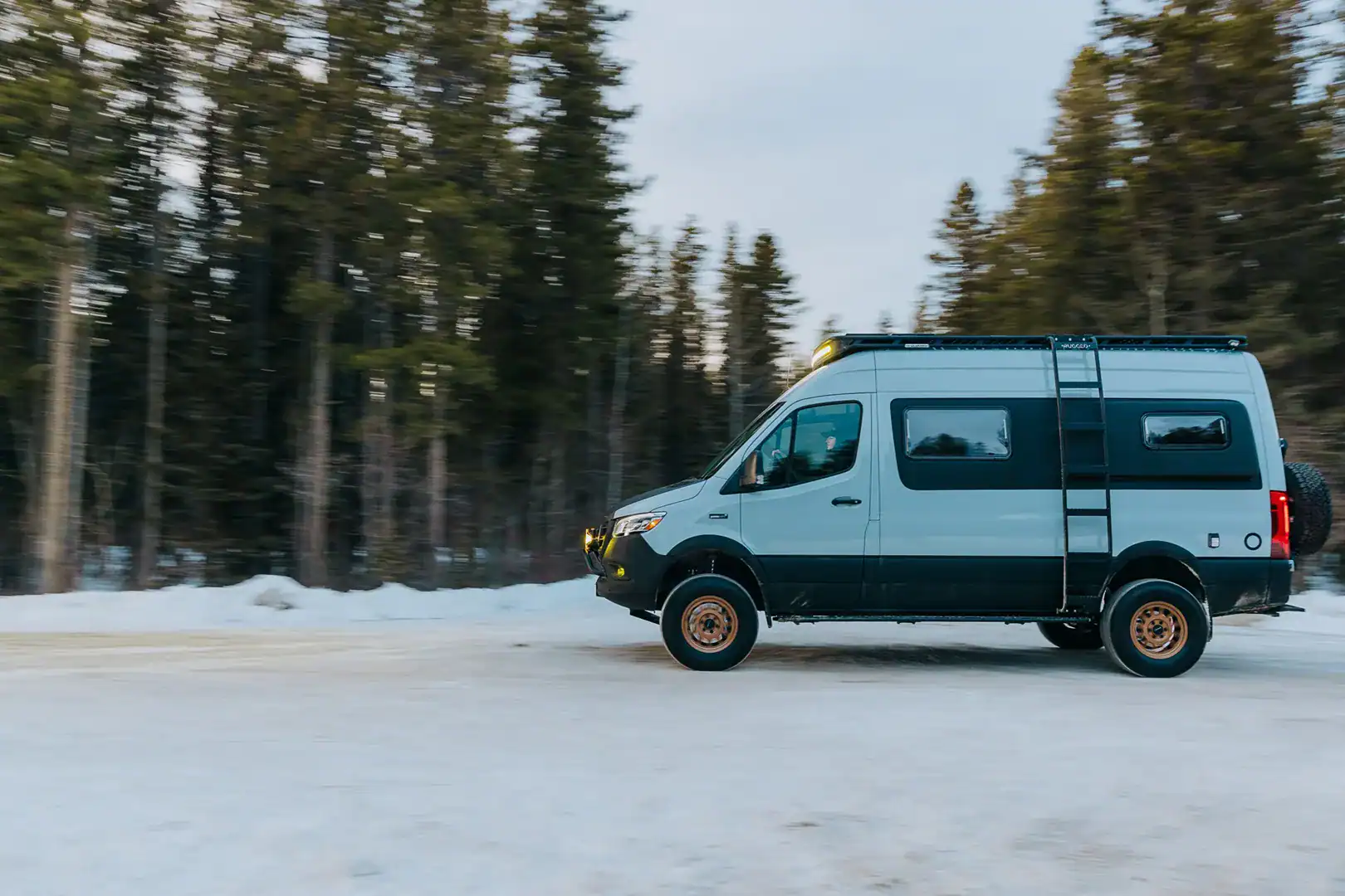 The Yama Vans Furō, a light blue Mercedes Sprinter 144 camper van with bronze wheels, driving on a snowy forest road