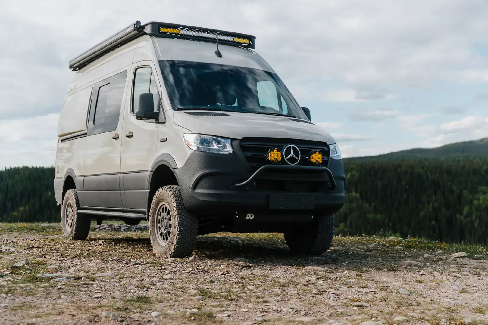 The Yama Vans Taiyō, a sand-colored Mercedes Sprinter 144 4x4 camper van with a roof light bar and amber lights, parked on a mountain overlook