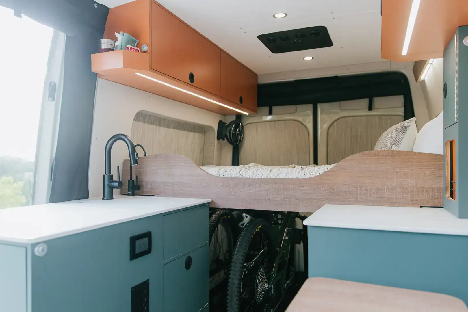 The Taiyō galley with a black faucet and teal cabinetry, with bikes stored in the gear garage beneath the bed