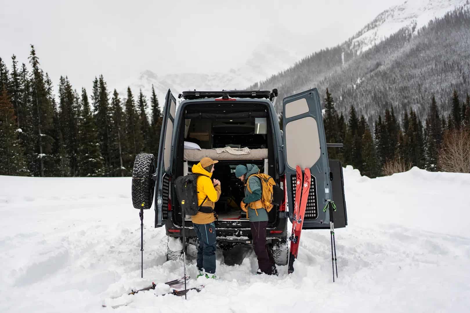 Two people loading skis and packs into the rear gear garage of a Yama Vans Teton 01N in the snow