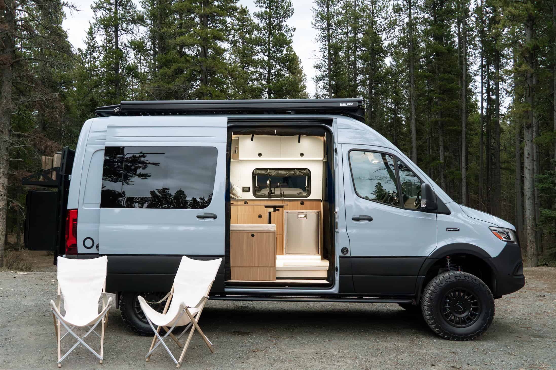 The Yama Vans Trekka, a light grey Mercedes Sprinter 144 camper van, parked in the forest with its sliding door open and camp chairs set out
