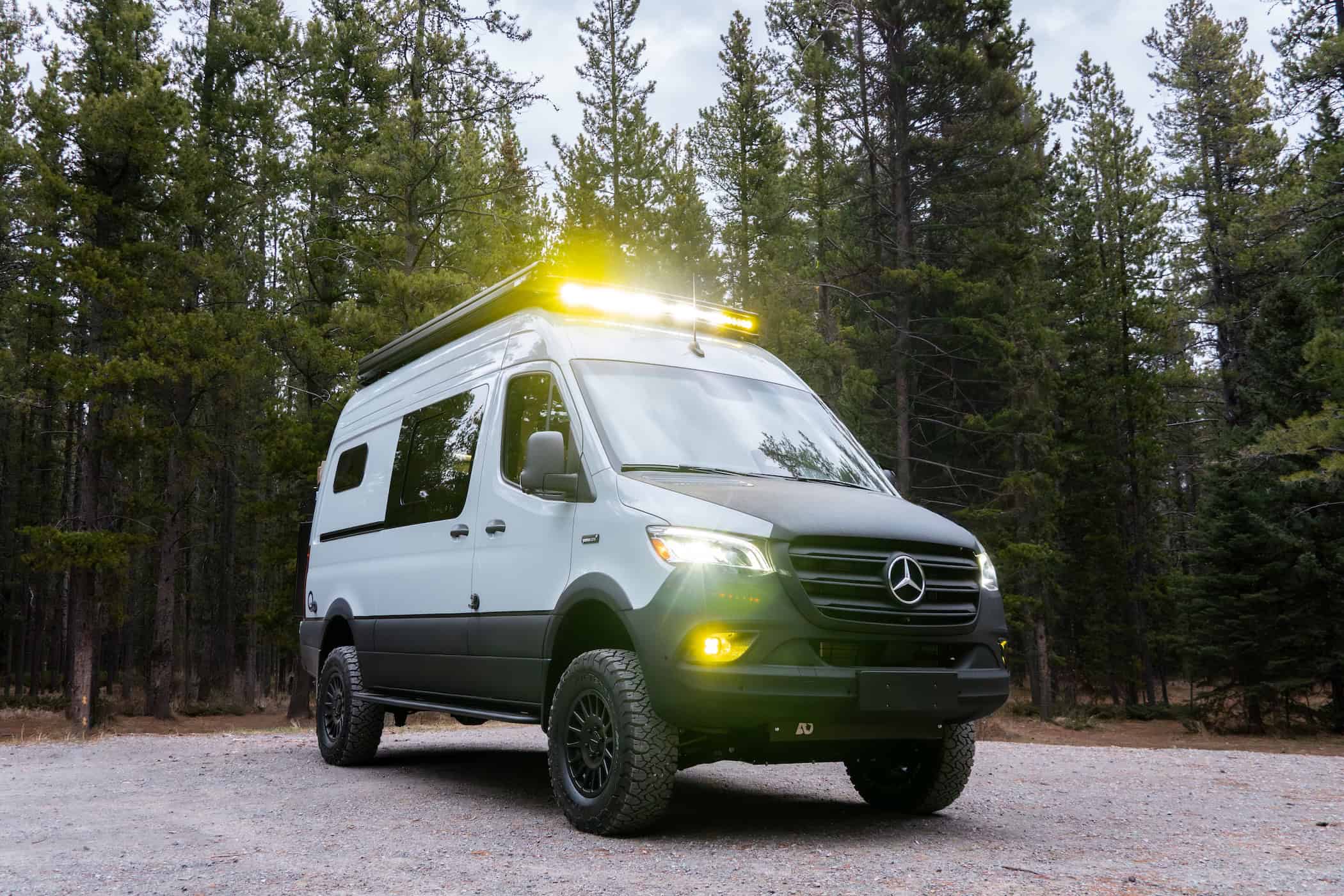 The front of the Yama Vans Trekka Sprinter 144 with off-road tires and a roof-mounted LED light bar in a pine forest