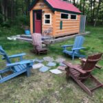 Small wooden tiny house with red roof in wooded area, outdoor seating.