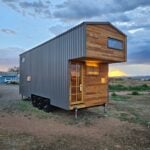 The Abundance of Less tiny house on a triple-axle trailer at sunset in Santa Fe, with cedar-clad cantilevered loft, grey metal siding, and a 12-pane entry door