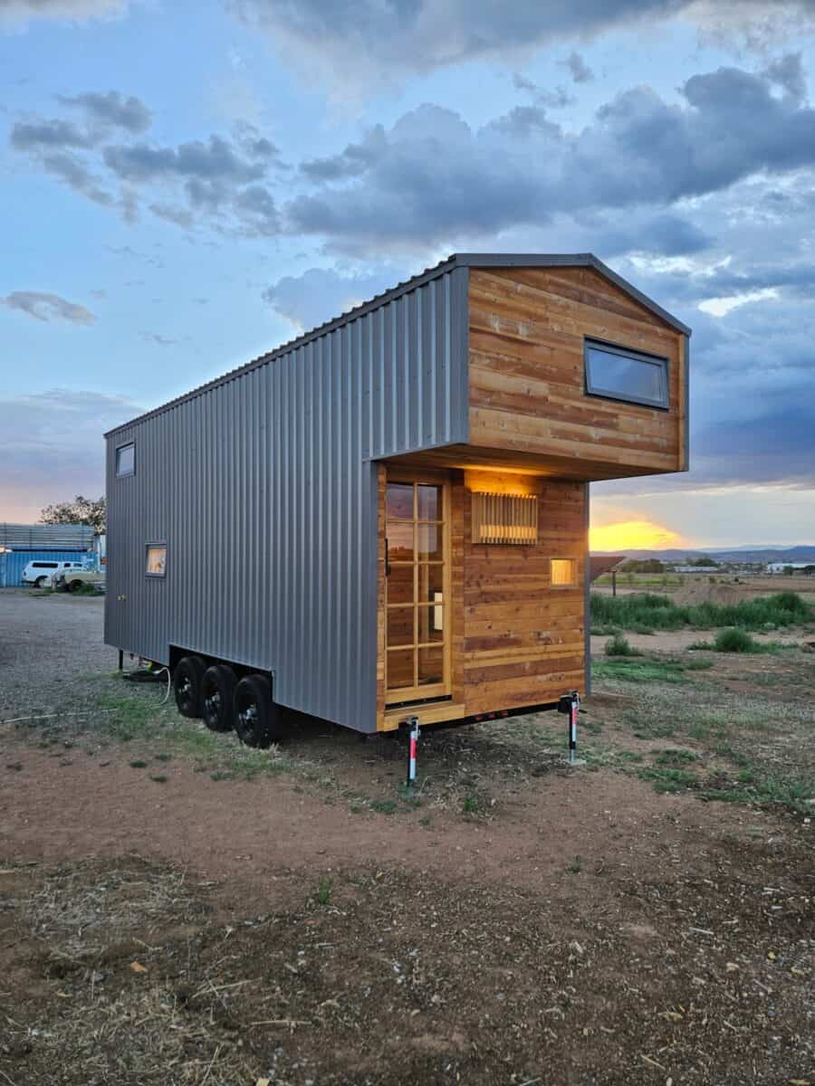The Abundance of Less tiny house on a triple-axle trailer at sunset in Santa Fe, with cedar-clad cantilevered loft, grey metal siding, and a 12-pane entry door