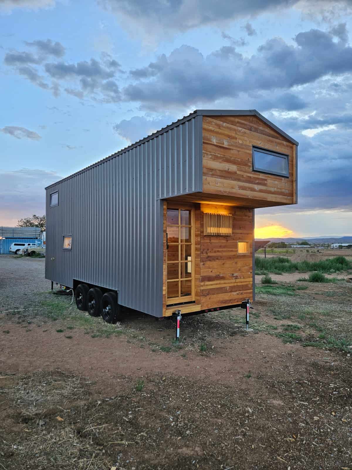 The Abundance of Less tiny house on a triple-axle trailer at sunset in Santa Fe, with cedar-clad cantilevered loft, grey metal siding, and a 12-pane entry door