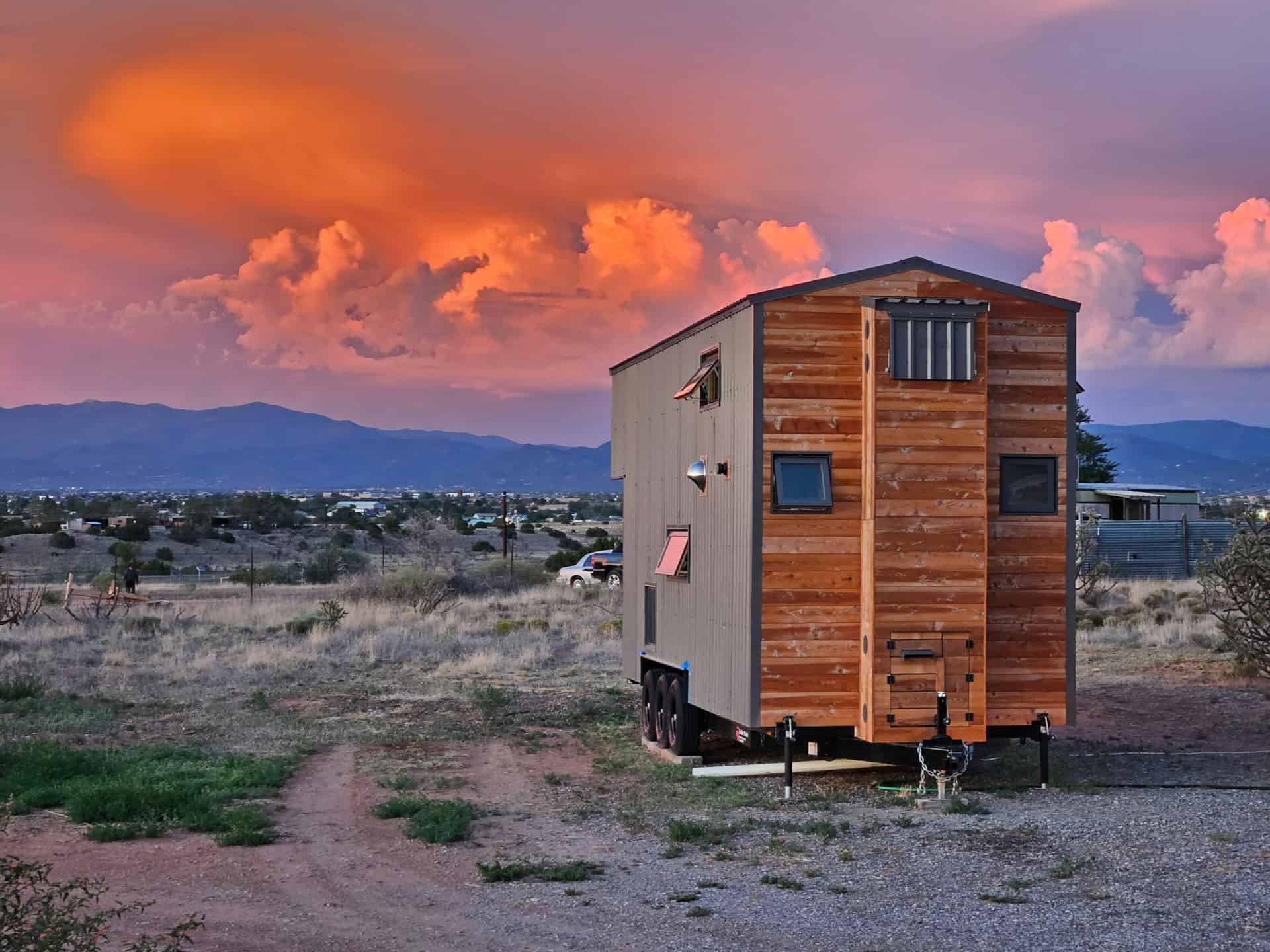 Rear gable of The Abundance of Less tiny house at sunset with wood cladding, open awning windows, triple-axle chassis, and mountains beyond Santa Fe