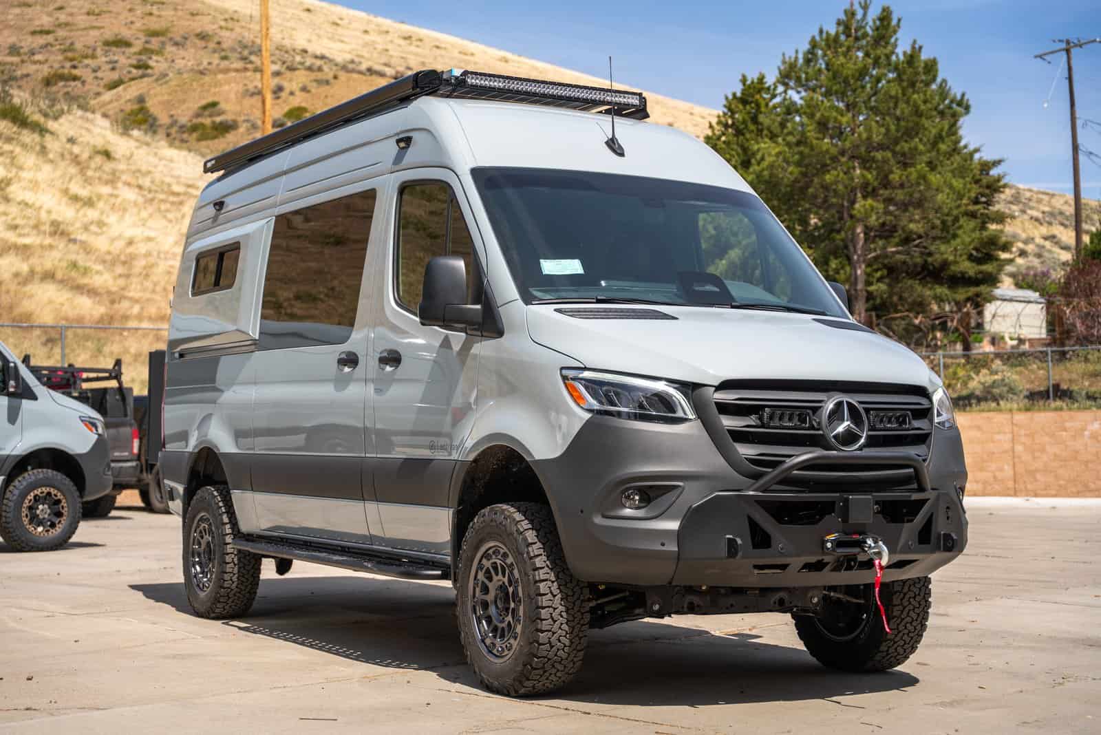 Silver Mercedes Sprinter 144 AWD camper van with a roof rack, ladder, black wheels, and a front winch