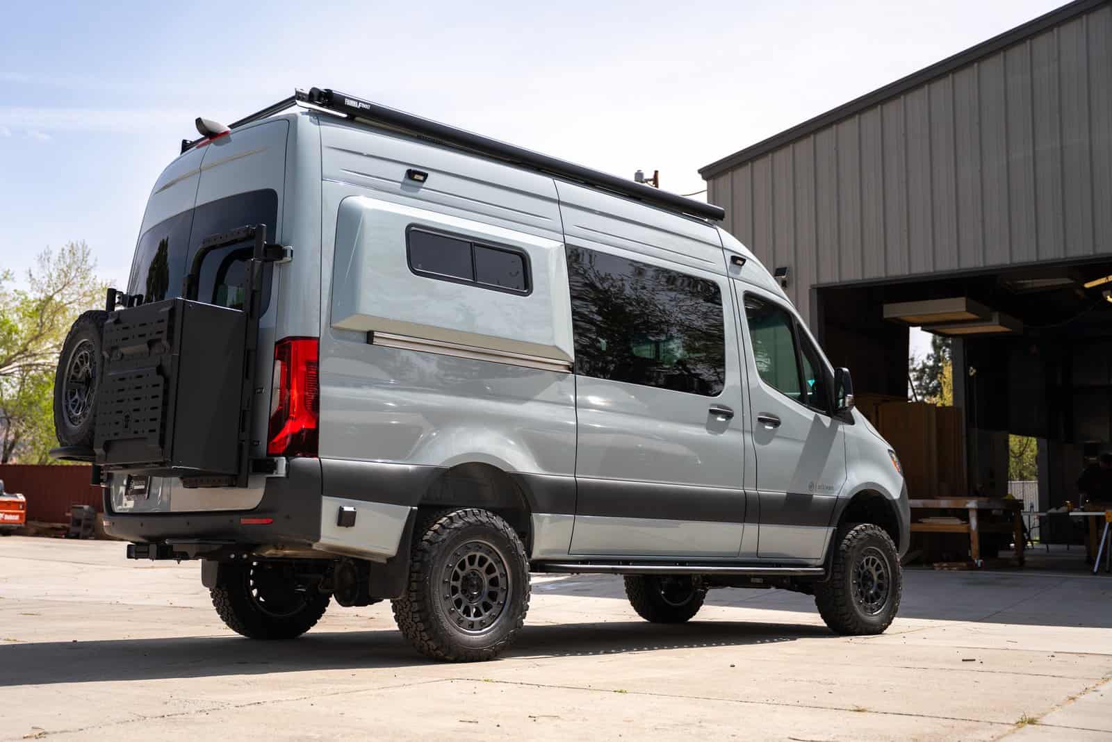 Rear three-quarter view of the silver Sprinter camper van with a rear-door spare tire carrier, ladder, and side window