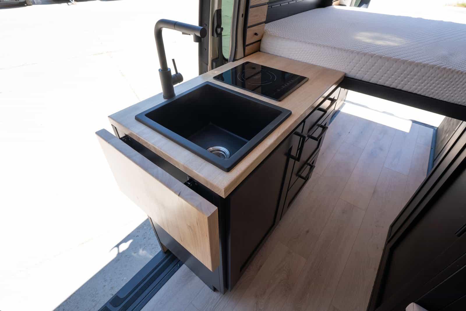 Galley kitchen with a black sink, induction cooktop, wood counters, and black cabinets next to the open sliding door